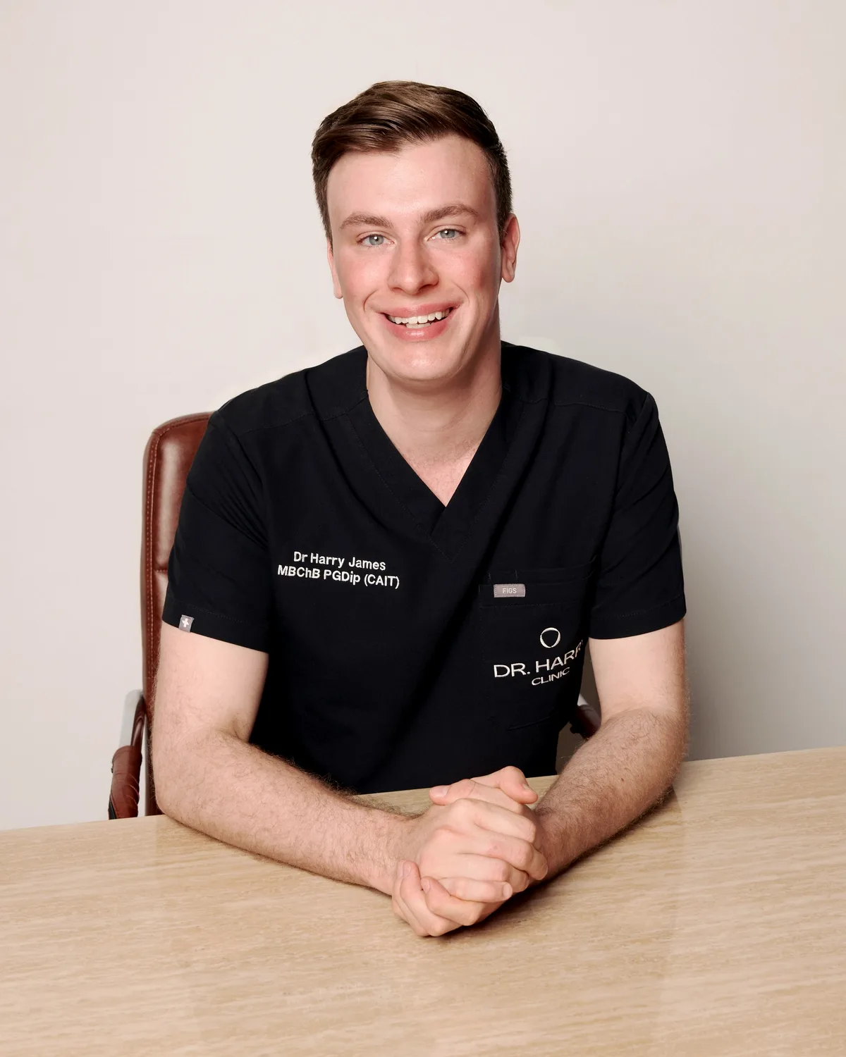 Dr Harry James smiling at the consultation desk in embroidered clinic scrubs, hands clasped in front of him.