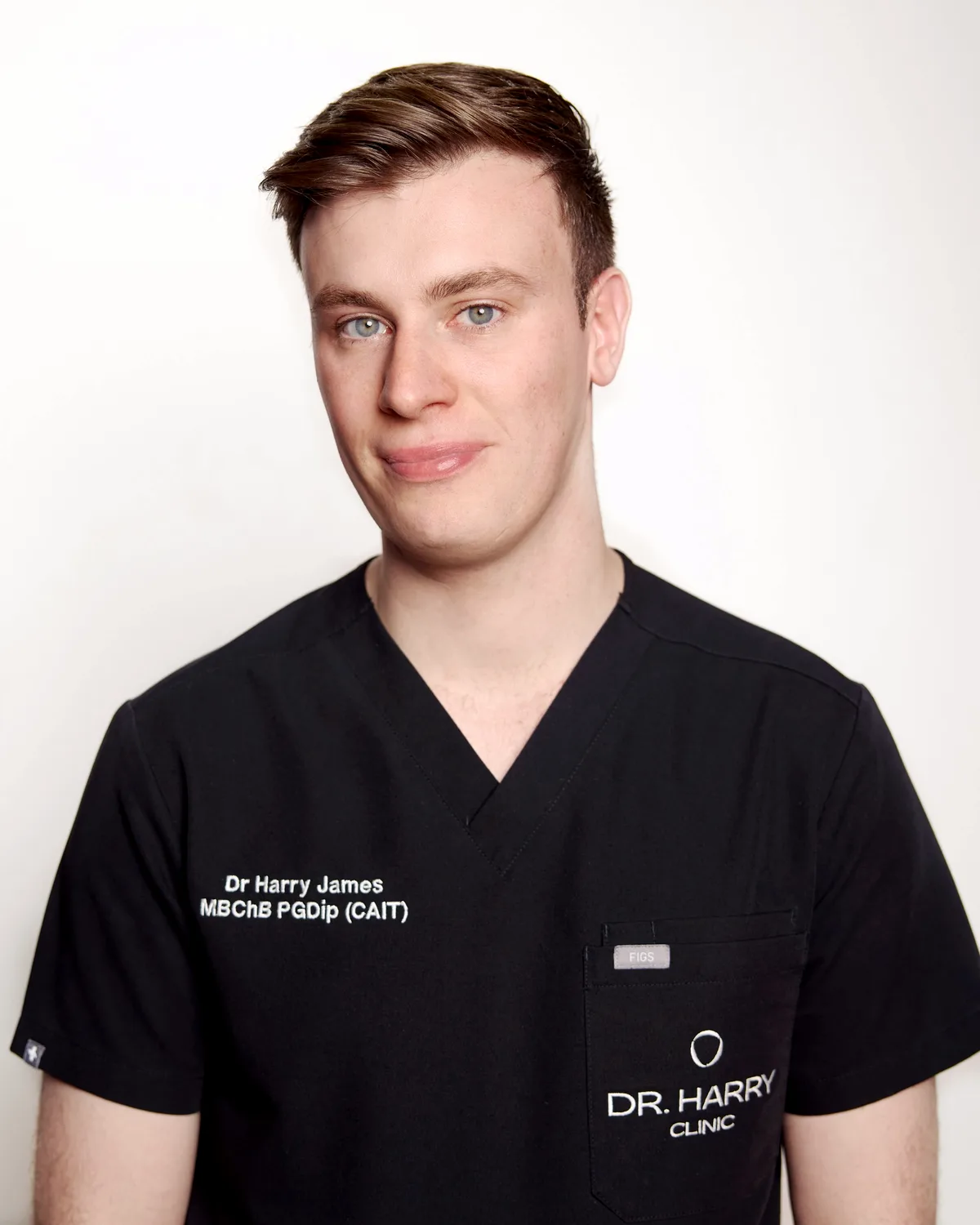 Studio portrait of Dr Harry James against a pale grey backdrop, smiling, in Dr Harry Clinic scrubs.