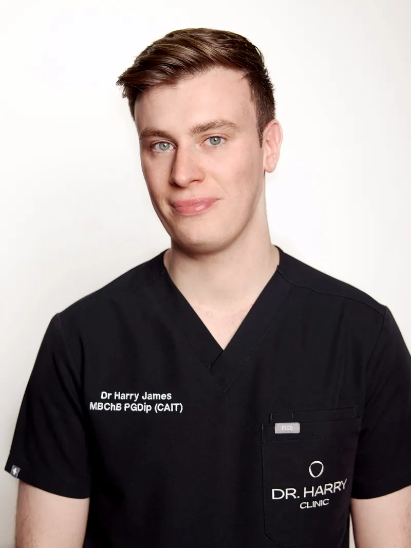 Studio portrait of Dr Harry James against a pale grey backdrop, smiling, in Dr Harry Clinic scrubs.