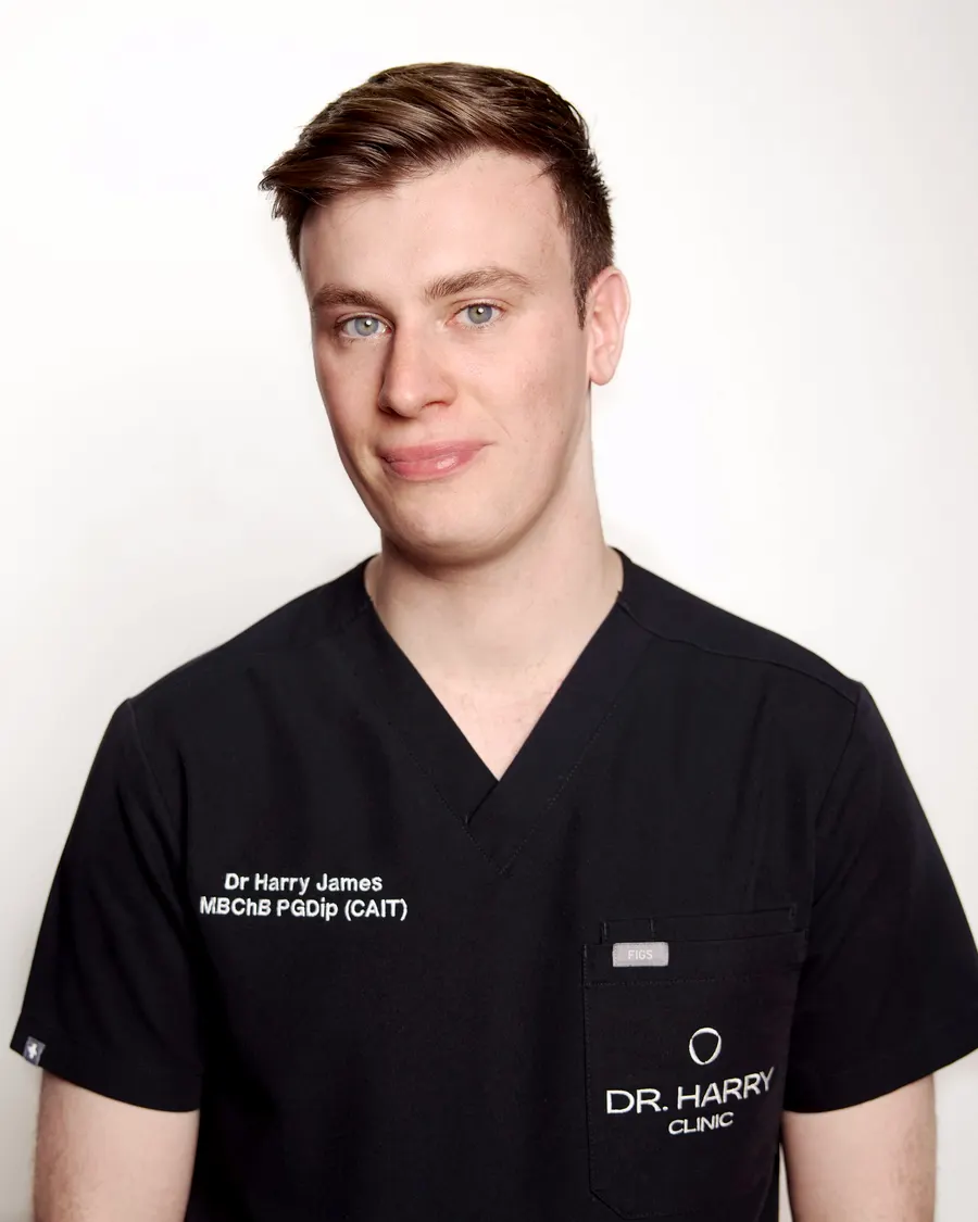 Studio portrait of Dr Harry James against a pale grey backdrop, smiling, in Dr Harry Clinic scrubs.