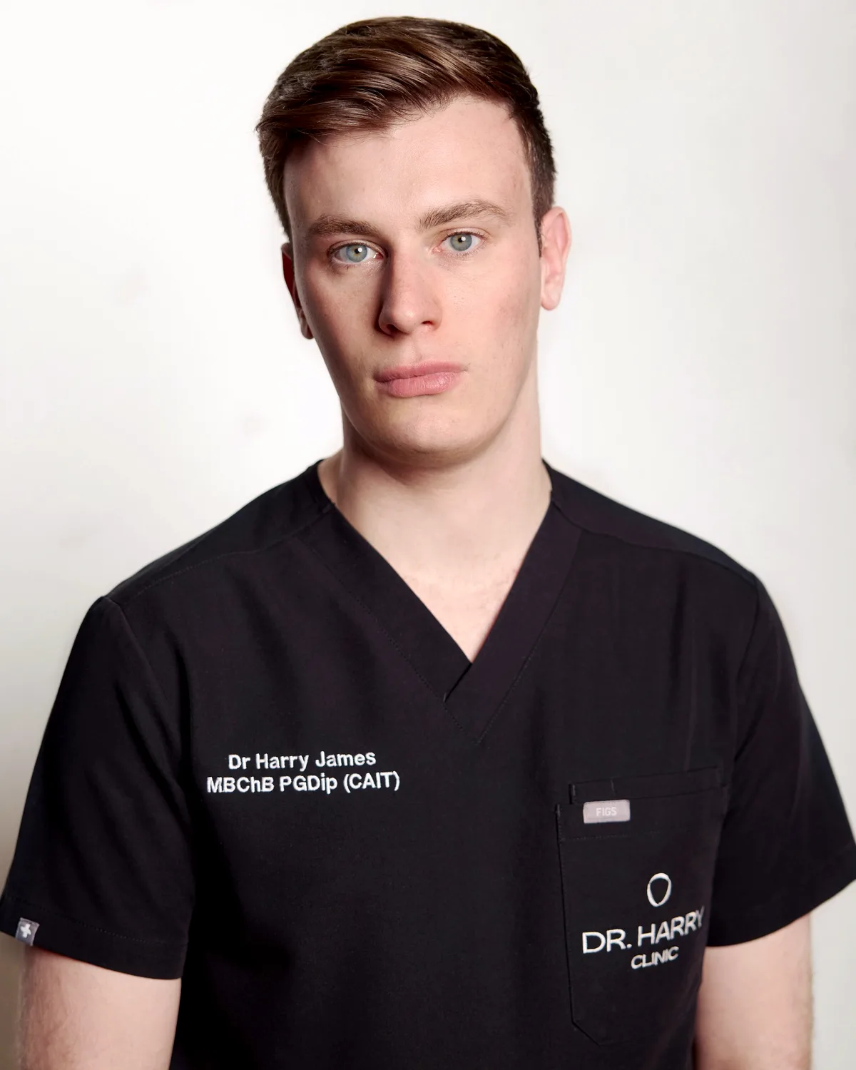 Studio portrait of Dr Harry James facing camera against a pale grey backdrop, scrubs embroidered Dr Harry James MBChB PGDip (CAIT).
