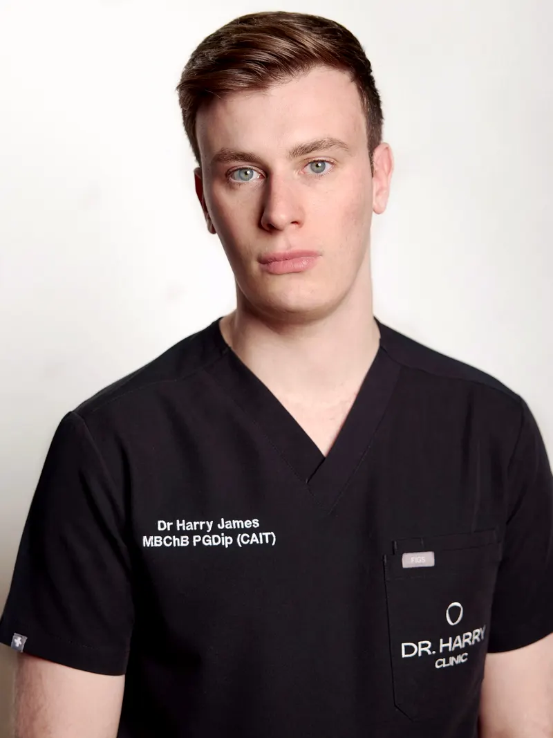 Studio portrait of Dr Harry James facing camera against a pale grey backdrop, scrubs embroidered Dr Harry James MBChB PGDip (CAIT).