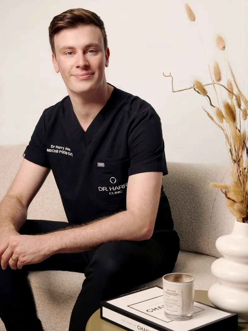 Dr Harry James seated on the clinic sofa in portrait crop, styled shelf and candle in the foreground.