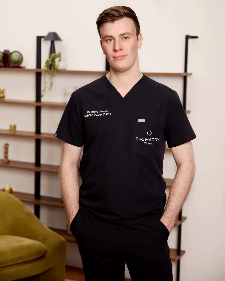 Dr Harry James standing in the consultation room, olive velvet armchair and styled shelving behind him.