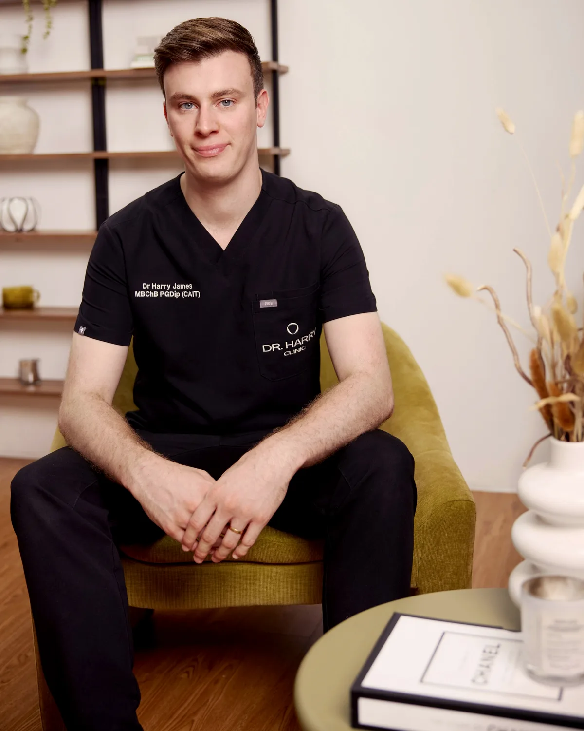 Dr Harry James seated in the olive velvet armchair, smiling, styled shelving behind him.