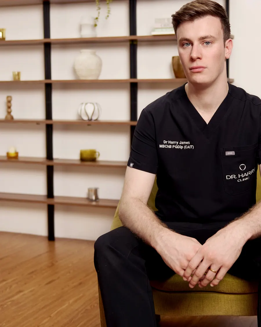 Dr Harry James seated in the olive armchair looking directly to camera, shelving and wood floor behind.