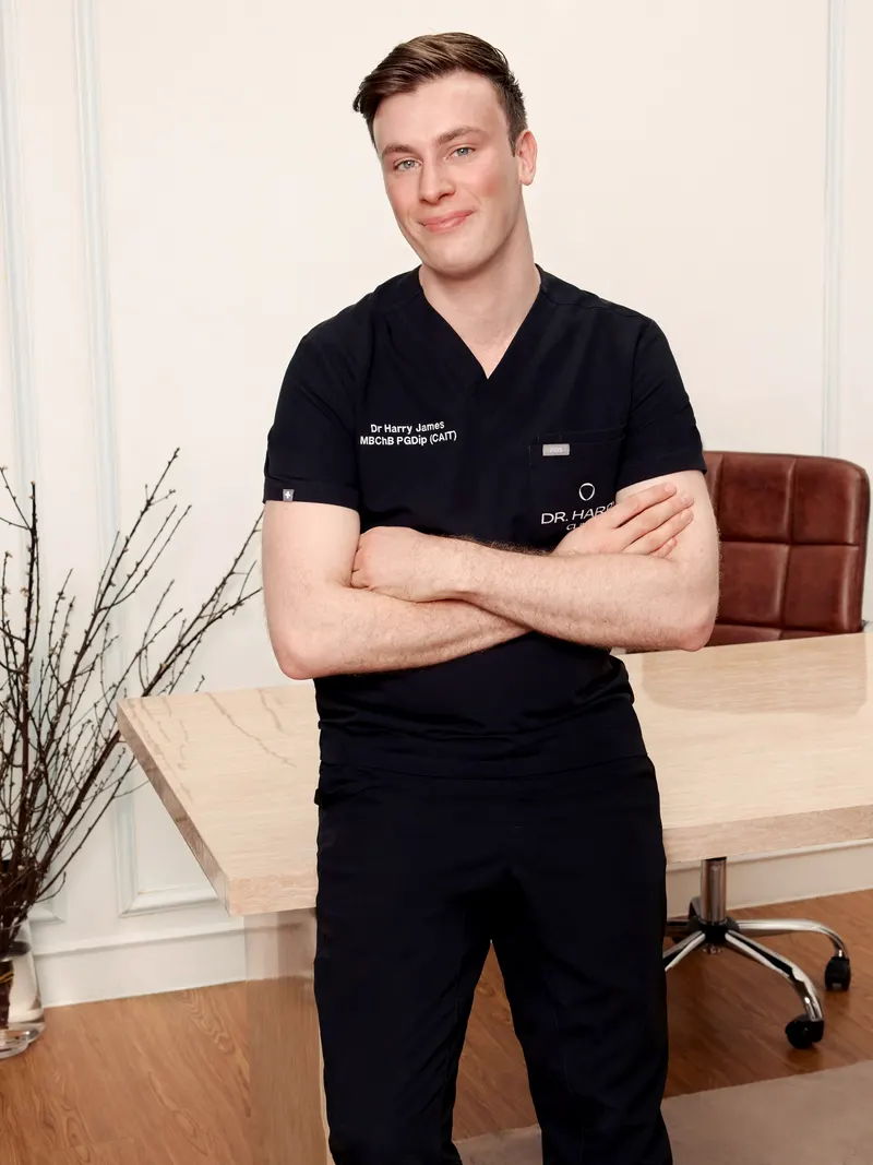 Dr Harry James leaning against the consultation desk with arms folded, panelled wall and wood flooring behind.