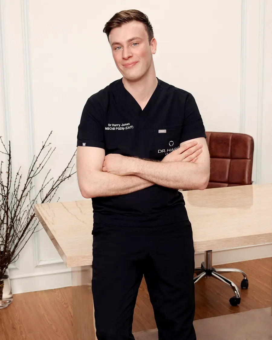 Dr Harry James leaning against the consultation desk with arms folded, panelled wall and wood flooring behind.