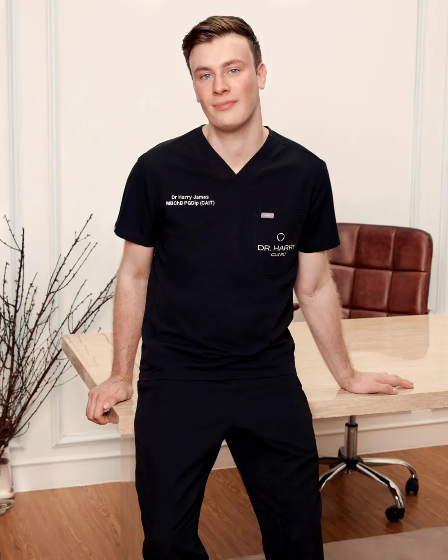 Dr Harry James resting one hand on the consultation desk, full length in black clinic scrubs.