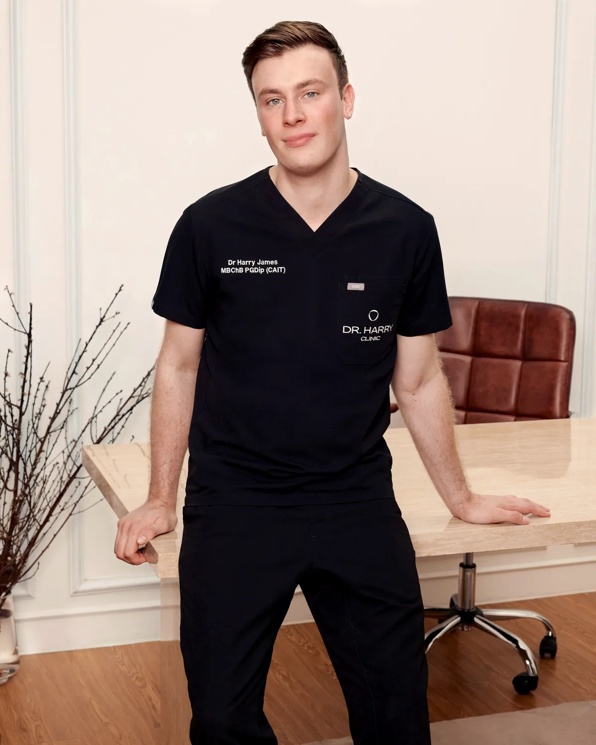 Dr Harry James resting one hand on the consultation desk, full length in black clinic scrubs.