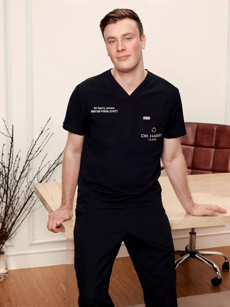 Dr Harry James resting one hand on the consultation desk, full length in black clinic scrubs.